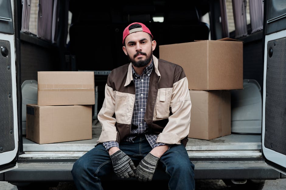 A young man with a beard, wearing a red cap backwards, a multi-colored jacket, a checkered shirt, dark jeans, and striped gloves, sitting on the open back of a white van. Behind him inside the van, there are several cardboard boxes of different sizes, some stacked on each other, and plastic containers, all used for packing belongings during a house relocation. The van is parked outdoors on a paved surface, with the vehicle's interior visible behind him. The scene suggests a furniture transport or packing process as part of home moving services provided by Man With a Van Edgware, which is involved in loading and moving household items. The lighting is natural, indicating daytime, and the overall environment reflects preparation for a moving day involving packing, loading, and transport of goods.