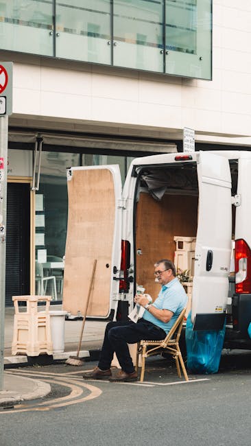 A man with glasses, wearing a light blue shirt and dark trousers, is seated on a wooden chair outside a white moving van with its rear doors open, which is positioned on a pavement in front of a modern building with large glass windows. Inside the van, visible packaging materials include cardboard boxes and wooden crates, some wrapped in bubble wrap. A wooden panel is propped against the side of the van, and a small stack of plastic crates is nearby. The scene depicts a home relocation process with furniture, boxes, and packing materials arranged for loading or unloading. The man appears to be taking a break during furniture transport or packing and moving activities, associated with house removals or relocation services, as provided by Man With a Van Edgware.