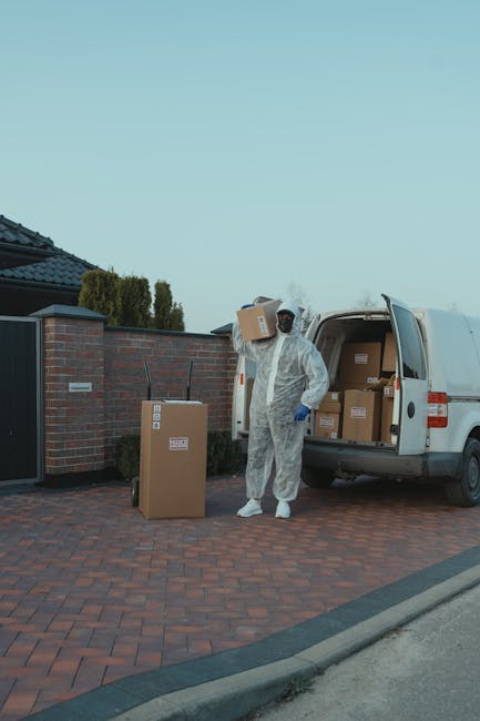 A young man with a beard, wearing a red cap backwards, a multi-colored jacket, a checkered shirt, dark jeans, and striped gloves, sitting on the open back of a white van. Behind him inside the van, there are several cardboard boxes of different sizes, some stacked on each other, and plastic containers, all used for packing belongings during a house relocation. The van is parked outdoors on a paved surface, with the vehicle's interior visible behind him. The scene suggests a furniture transport or packing process as part of home moving services provided by Man With a Van Edgware, which is involved in loading and moving household items. The lighting is natural, indicating daytime, and the overall environment reflects preparation for a moving day involving packing, loading, and transport of goods.