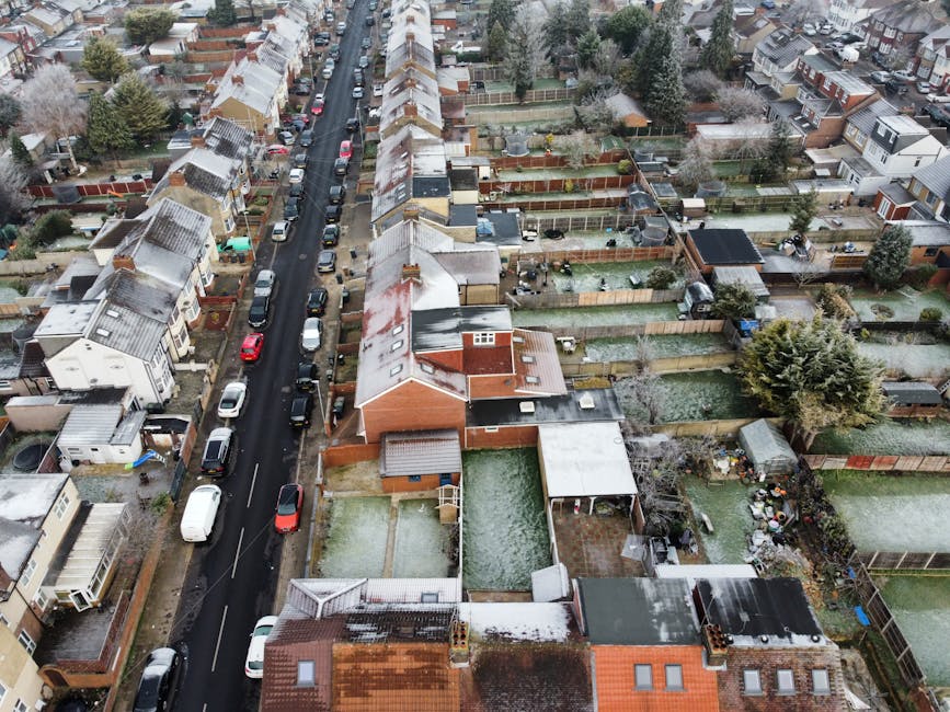 An aerial view of a residential street during winter showing rows of terraced houses with snow-covered roofs and parked cars lining the narrow asphalt road. Behind the houses, there are private gardens enclosed by wooden fences, some with sheds, patio furniture, and outdoor storage. Several properties have backyards with visible paving, lawn areas, and garden features. A van with a sliding side door is parked on the pavement near one of the houses, indicating a home relocation or furniture transport process underway. The van appears to be part of Man With a Van Edgware's moving fleet, supporting house removals and packing and moving services in the Hale Lane area of Edgware. The scene is evenly lit, typical of overcast winter weather, highlighting the organized arrangement of residential properties, vehicles, and outdoor spaces, reflecting a typical neighborhood landscape prepared for a professional moving or home relocation service.