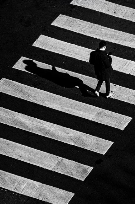 A black and white aerial photograph shows a man crossing a zebra crossing on a paved road. He is wearing a dark suit and light-colored shoes, and is holding a mobile phone in his right hand. The pedestrian crossing consists of alternating white and dark stripes, with the white stripes being textured and the dark stripes appearing smooth. The man's shadow is cast on the crossing, indicating a strong light source from the upper left side of the image. The scene captures the moment during the day, emphasizing urban movement and pedestrian activity, relevant to house removals and the logistical aspects of home relocation within an urban environment, as discussed on the Edgware removals guide page for Hale Lane homes.