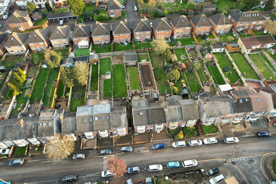 An aerial view of a residential street during winter showing rows of terraced houses with snow-covered roofs and parked cars lining the narrow asphalt road. Behind the houses, there are private gardens enclosed by wooden fences, some with sheds, patio furniture, and outdoor storage. Several properties have backyards with visible paving, lawn areas, and garden features. A van with a sliding side door is parked on the pavement near one of the houses, indicating a home relocation or furniture transport process underway. The van appears to be part of Man With a Van Edgware's moving fleet, supporting house removals and packing and moving services in the Hale Lane area of Edgware. The scene is evenly lit, typical of overcast winter weather, highlighting the organized arrangement of residential properties, vehicles, and outdoor spaces, reflecting a typical neighborhood landscape prepared for a professional moving or home relocation service.