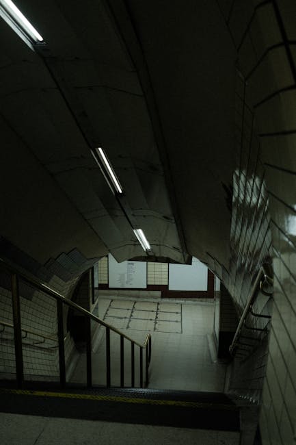 A downward view of the staircase and underground passageway at Edgware Underground Station, featuring tiled walls and ceiling, metal handrails, and fluorescent lighting. The tiled flooring extends from the staircase to a tiled platform area. This setting is part of a typical station environment, with no furniture or moving equipment visible. The image captures a quiet, well-lit area suitable for access advice related to station household relocations or furniture transport near the station, with the accompanying professional removals service provider, Man With a Van Edgware, supporting efficient and careful home relocation and moving logistics.