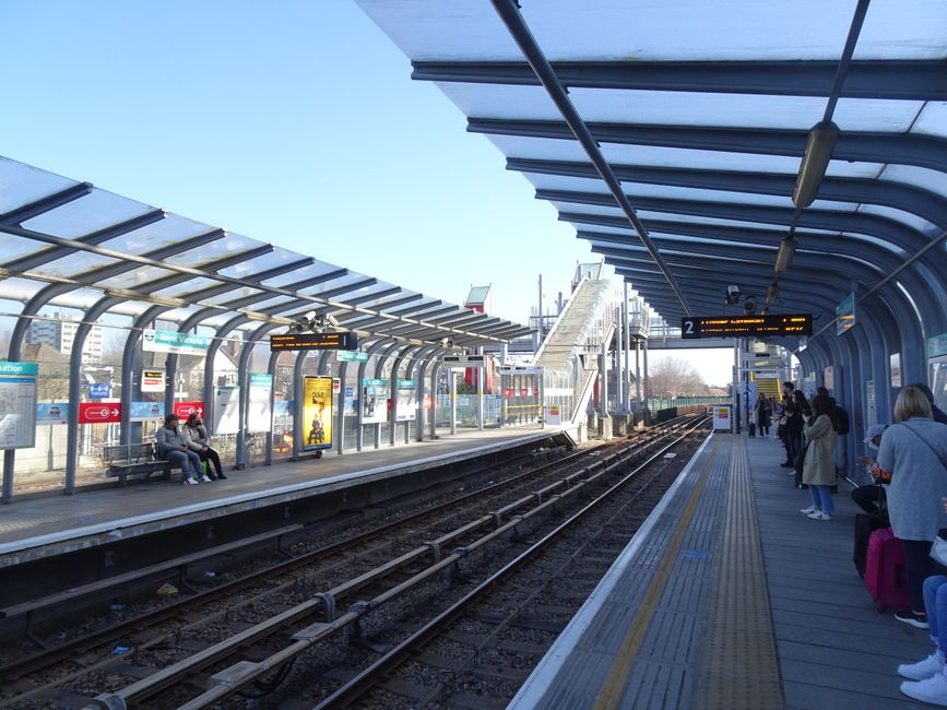 The image depicts a train station platform at Edgware with a curved, transparent roof structure supported by metal beams. On the left side, there are two people sitting on a bench beneath a shelter, surrounded by advertisements and station signage. In the background, a train is approaching or waiting at the platform, with station stairs and additional signage visible. Inside the station, on the right side of the platform, several passengers are standing or sitting, some with luggage or backpacks, awaiting their train. The platform features tactile paving strips for accessibility, and the environment is well-lit with natural daylight, highlighting the modern, clean design of the station. This setting reflects typical urban railway infrastructure, suitable for inclusion in content about house removals or moving logistics when considering commute arrangements or transport access for relocation services managed by Man With a Van Edgware.