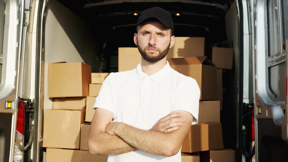 A young man with a serious expression, wearing a black cap and white polo shirt, stands inside an open van loaded with cardboard boxes of various sizes, some wrapped in plastic or packing materials. T