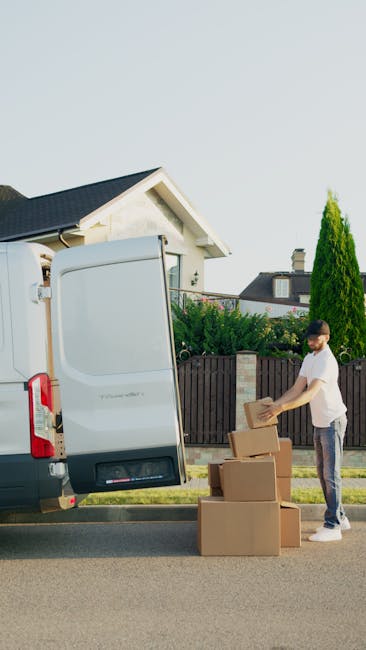 A young man with a serious expression, wearing a black cap and white polo shirt, stands inside an open van loaded with cardboard boxes of various sizes, some wrapped in plastic or packing materials. T